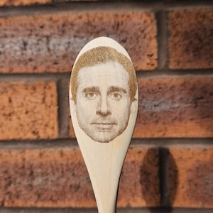 Puede incluir: Una cuchara de madera con un retrato del rostro de un hombre grabado en la cuchara. El retrato es de color marrón claro y la cuchara está hecha de madera clara. El fondo es una pared de ladrillos.