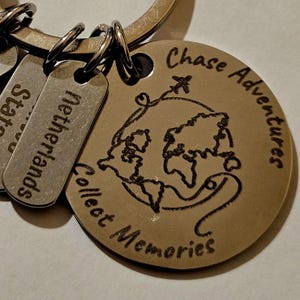 May include: A silver keyring featuring a round charm engraved with a world map and the words "Chase Adventures Collect Memories." Rectangular tags are engraved with the word "Netherlands" and "States".