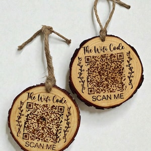 May include: Two wooden WiFi code signs, each cut from a tree slice. The signs have a QR code, the text "The Wifi Code" and "SCAN ME" engraved on them. Each sign has a twine loop for hanging.
