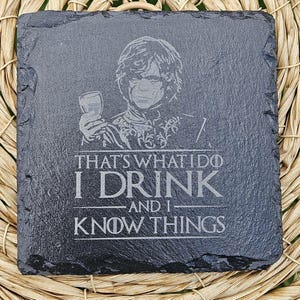 Könnte beinhalten: Quadratischer, dunkelgrauer Schieferuntersetzer mit einem geätzten Bild einer Person, die ein Glas hält, und dem Text "THAT'S WHAT I DO I DRINK AND I KNOW THINGS". Der Untersetzer liegt auf einer hellbraunen, gewebten Oberfläche.
