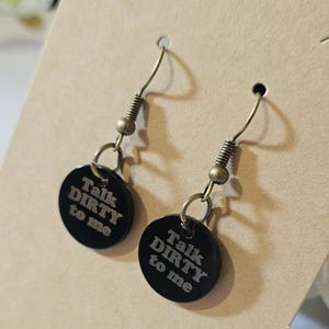 May include: A pair of black disc earrings with the text "Talk DIRTY to me" in a white, sans-serif font. The earrings have bronze-colored hooks and springs. The earrings are displayed on a tan card.