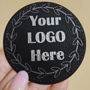 May include: A black, round coaster with the text "Your LOGO Here" in silver lettering. A white, leaf-like design encircles the text. The coaster is held by a person.