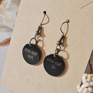 May include: A pair of black disc earrings with the text "one ear out" and "one ear in". The earrings have a hook and spring design with a bronze finish. The earrings are displayed on a tan card.
