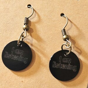 May include: A pair of black disc earrings with the text "I am listening" engraved on each. The earrings have bronze-colored hooks, springs, and beads. The earrings are displayed on a tan surface.