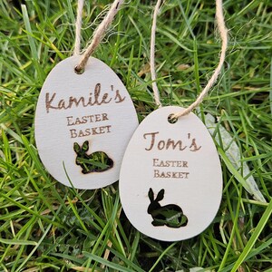 May include: Two wooden Easter basket tags shaped like eggs with bunny cutouts. One tag says "Kamile's Easter Basket" and the other says "Tom's Easter Basket".
