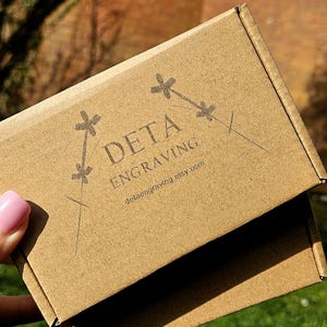 Puede incluir: Una caja de cartón marrón con el texto "DETA ENGRAVING" y la dirección del sitio web detaengraving.etsy.com. La caja tiene un diseño decorativo de cruces y líneas. La caja es sostenida por una persona con esmalte de uñas rosa.