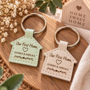 Pode incluir: Dois porta-chaves em forma de casa com argolas de metal, um em verde sálvia e outro em bege. Cada um apresenta o texto gravado "Our First Home", um ícone de coração, os nomes "SYDNEY & AMELKA" e um motivo de ramo com um pequeno coração.