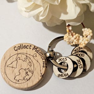 May include: A wooden keyring featuring a round charm engraved with the words "Collect Memories" and a world map design. It has smaller wooden circles with dates and the word "INDIA". The keyring is silver.