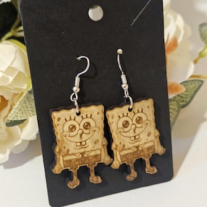 May include: Wooden dangle earrings featuring a cartoon character design. The earrings are light brown with a detailed engraving of a smiling character. The earrings hang from silver-colored fish hook ear wires. The earrings are displayed on a black card.