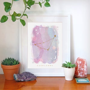 May include: A framed print with a watercolor background in shades of pink, purple, and blue. The constellation Capricorn is depicted in gold with a gold star at the top. The text "CAPRICORN" is printed in gold at the bottom of the print.