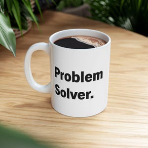 May include: White ceramic coffee mug with a black handle. The mug has black text that reads "Problem Solver."