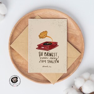 May include: A greeting card with a vintage gramophone illustration in red and gold, with the German text "Du bringst mein Herz zum Tanzen" (You make my heart dance). The card is on a wooden plate with a brown envelope.