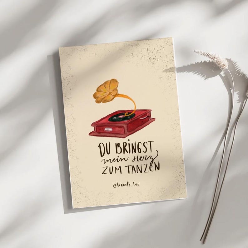 May include: A greeting card with a vintage gramophone illustration in red and gold. The card features the German phrase "DU BRINGST mein HERZ ZUM TANZEN" in black script, with the artist's name, "@beats_leo," below.