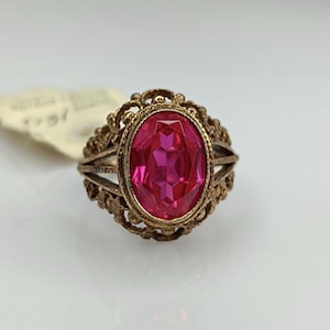 May include: A vintage gold-toned ring featuring an oval, faceted, bright pink gemstone. The ring has an ornate filigree design around the gemstone and on the band. The ring is a classic piece of jewelry.
