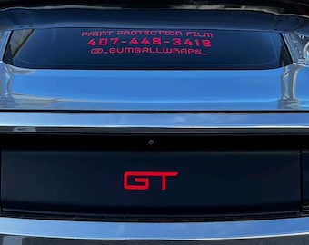 May include: A silver car with a large black rear spoiler featuring NRG branding. The rear window displays red text reading PAINT PROTECTION FILM 407-448-3418 @_GUMBALLWRAPS_. The trunk panel features a red GT emblem between red vertical taillights.