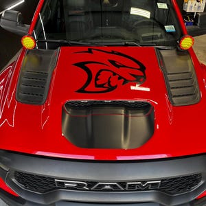 May include: A bright red pickup truck featuring a prominent black Hellcat logo decal centered on the hood. The vehicle includes a large black hood scoop, black side vents, a black front grille with RAM lettering, and two round yellow auxiliary lights.