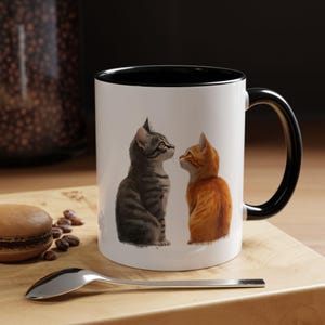 May include: White ceramic mug with a black rim and handle. The mug features a graphic of two cats looking at each other. The cat on the left is gray and the cat on the right is orange.
