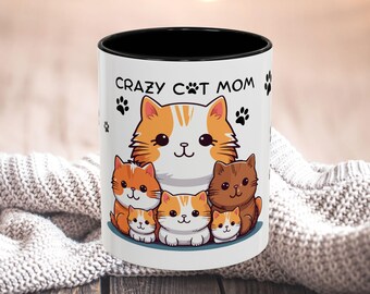 Könnte beinhalten: Weiße Keramiktasse mit schwarzem Rand und Henkel. Die Tasse zeigt eine Comic-Illustration einer Katzenmutter und ihrer Kätzchen. Der Text "Crazy Cat Mom" ist auf die Tasse gedruckt.