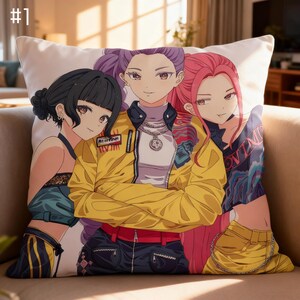 Puede incluir: Cojín cuadrado blanco con una colorida ilustración de estilo anime de tres mujeres jóvenes. La figura central lleva una chaqueta amarilla, mientras que las otras visten atuendos azules y negros. El cojín está sobre un sofá beige.