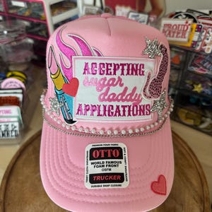 May include: Pink trucker hat with the text "ACCEPTING sugar daddy APPLICATIONS". The hat features a lipstick and heart graphic, a pearl chain, and a leopard print design. The hat has a snap closure.