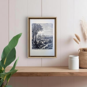 Op de afbeelding: Een ingelijste zwart-wit landschapsprint met een gedetailleerde illustratie van een stadsgezicht met hoge torengebouwen, bomen en een rivier. De print zit in een goudkleurige lijst en staat op een houten plank.