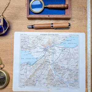 Op de afbeelding: Een assortiment vintage items op een houten oppervlak. Inclusief een houten vergrootglas in een doos, een houten pen, een kaart met de tekst "ENVIRONS D'INTERLAKEN", een kompas, een liniaal en een blauwe glazen bol.