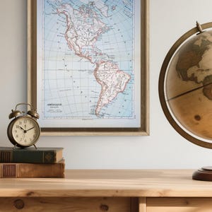Peut inclure: Une carte vintage encadrée des Amériques, avec le mot "AMÉRIQUE" en bas. La carte est dans un cadre en bois clair, et se trouve sur un bureau en bois avec un globe et un réveil. La carte montre les continents en nuances de bleu, beige et rouge.