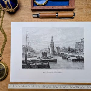 May include: A vintage brass compass on a chain, a wooden pen, and a framed magnifying glass are on a wooden surface. A black and white print of a European cityscape with boats and a tall tower is also present.
