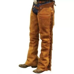 Op de afbeelding: Bruine leren chaps met franjes en een riem met een zilveren gesp. De chaps worden over een blauwe jeans gedragen.