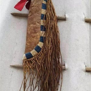 Könnte beinhalten: Eine handgefertigte Lederscheide mit einem blau-beigen Perlendesign, verziert mit langen, ausgefransten Strähnen. Ein rotes Stoffstück und ein Holzgriff sind oben befestigt. Der Artikel hängt vor einem weißen Hintergrund.