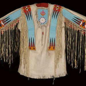 Peut inclure: Une chemise en cuir blanc avec des motifs perlés en bleu, rouge, orange et noir. La chemise a des franges autour des bords et un motif perlé sur la poitrine.