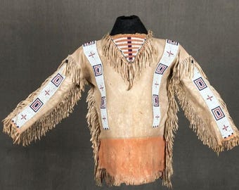 Puede incluir: Una camisa de ante de color canela con flecos y abalorios decorativos. La camisa presenta rayas de cuentas blancas con diseños geométricos y cruces, un cuello negro y una sección inferior naranja. Las mangas y los hombros están adornados con flecos.
