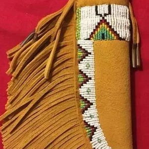 Könnte beinhalten: Senfgelbe Leder-Messerscheide mit einem Perlendesign in Weiß, Grün, Rot und Braun. Die Scheide hat Fransen und einen Griff. Der Messergriff ist sichtbar.