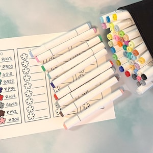 May include: A collection of colorful art markers with white barrels and color-coded tips, stored in a black case. A color chart with flower-shaped icons and color codes like B003, R503, and G318 is also present, suggesting a creative art supply set.