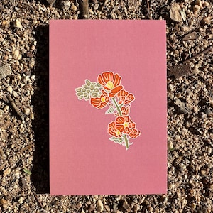 May include: A pink greeting card featuring a floral illustration. The illustration depicts orange flowers with yellow centers and green leaves. The card is rectangular and rests on a bed of small rocks and pebbles.