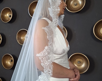 May include: A white wedding veil with intricate lace detailing along the edges. The veil is long and flowing, and the lace is embellished with sequins. The bride is wearing a white dress. The background is a dark wall with gold accents.