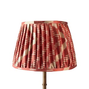 May include: A pleated lampshade with a coral and cream ikat pattern. The lampshade has a tapered shape and is mounted on a brass-coloured metal stand. The fabric has a textured appearance, and the design features horizontal and vertical lines.