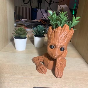 May include: A brown, wooden, Groot-shaped planter with a smiling face. The planter is holding a small green plant. There are two other small green plants in white pots on a wooden surface.