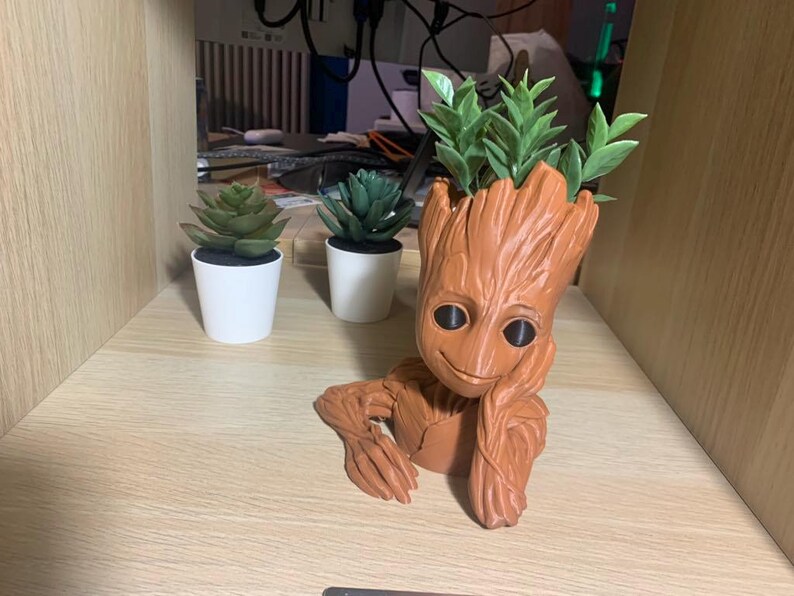 May include: A brown, wooden, Groot-shaped planter with a smiling face. The planter is holding a small green plant. There are two other small green plants in white pots on a wooden surface.
