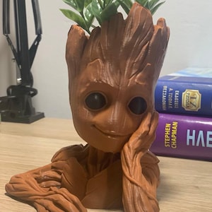 May include: A brown 3D printed planter shaped like the character Groot from the Guardians of the Galaxy movie series. The planter has a smiling face and is holding a hand to his chin. A small green plant is growing out of the top of the planter.