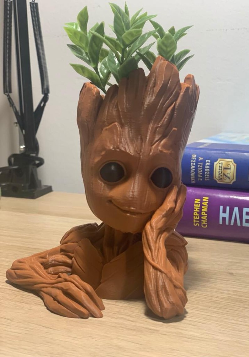 May include: A brown 3D printed planter shaped like the character Groot from the Guardians of the Galaxy movie series. The planter has a smiling face and is holding a hand to his chin. A small green plant is growing out of the top of the planter.