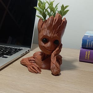 May include: A terracotta-coloured Groot planter with a smiling face, holding a small plant with green leaves. The planter is sitting on a wooden surface.