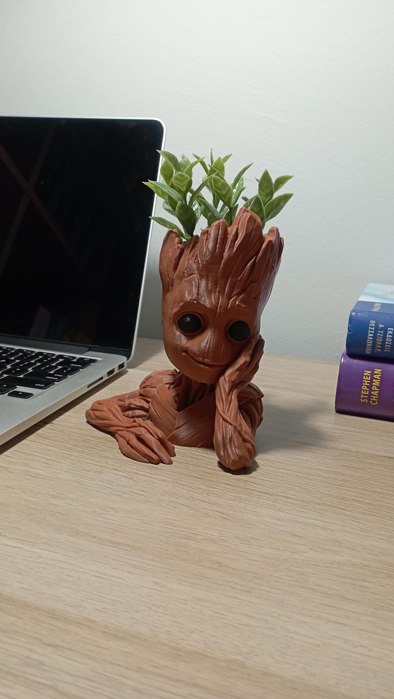 May include: A terracotta-coloured Groot planter with a smiling face, holding a small plant with green leaves. The planter is sitting on a wooden surface.