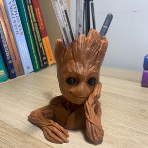 May include: A brown, 3D printed Groot figurine with a smiling face and a pen holder in the top of its head. The figurine is sitting on a wooden surface with several books in the background.