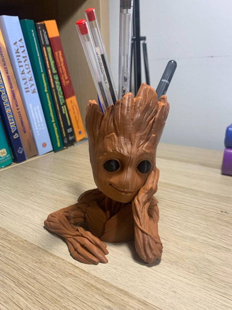 May include: A brown, 3D printed Groot figurine with a smiling face and a pen holder in the top of its head. The figurine is sitting on a wooden surface with several books in the background.