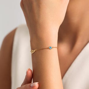 May include: A delicate gold bracelet with a small, openwork wing charm and a round, light blue gemstone. The bracelet is worn on a wrist, highlighting its simple yet elegant design. The jewellery is a subtle accessory.