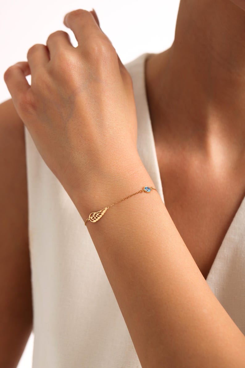 May include: A delicate gold bracelet with a wing charm and a small blue gemstone. The bracelet is worn on a wrist, highlighting its fine details and elegant design. The jewelry is a subtle accessory.