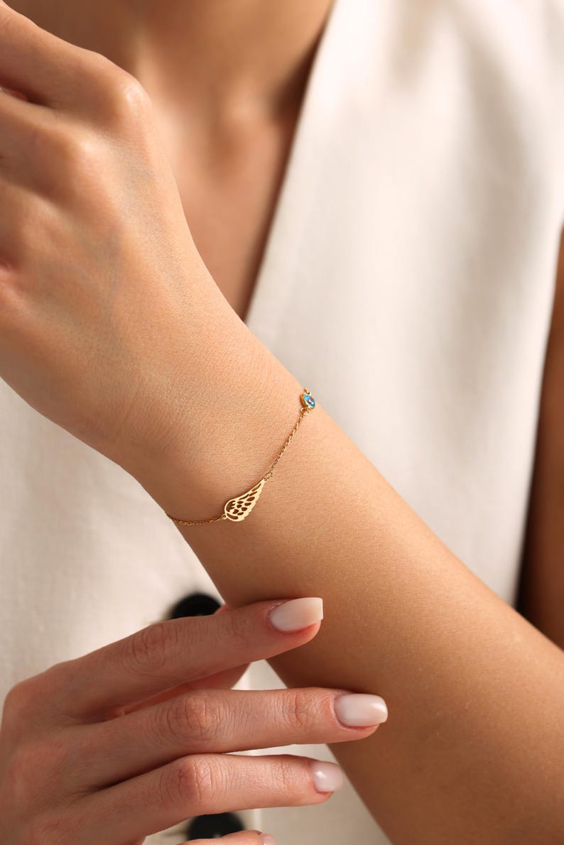 May include: A delicate gold bracelet featuring a wing charm and a small blue gemstone. The bracelet is worn on a wrist, showcasing its fine details and elegant design. The jewelry is a stylish accessory.
