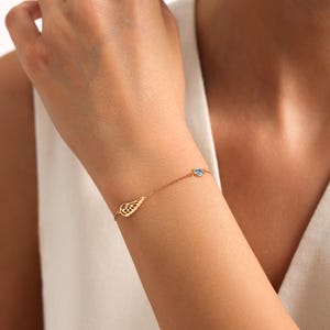 May include: A delicate gold bracelet with a wing charm and a small blue gemstone. The bracelet is worn on a wrist, highlighting its fine details and elegant design. The jewelry is a subtle accessory.