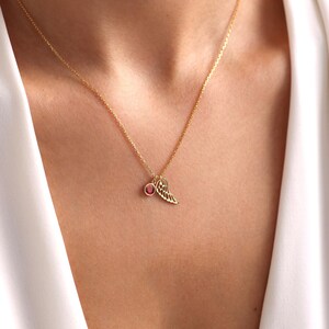 Op de afbeelding: Een gouden ketting met een kleine rode edelsteen en een delicate gouden vleugelbedel.